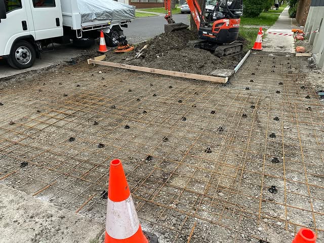 Steel mesh and bar chairs set out for a driveway pour, excavator behind