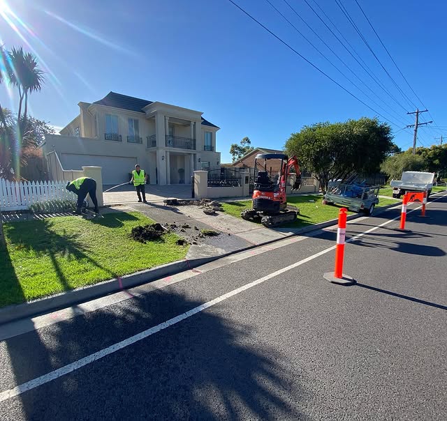 Crew with a mini excavator digging along a nature strip, cones on the road