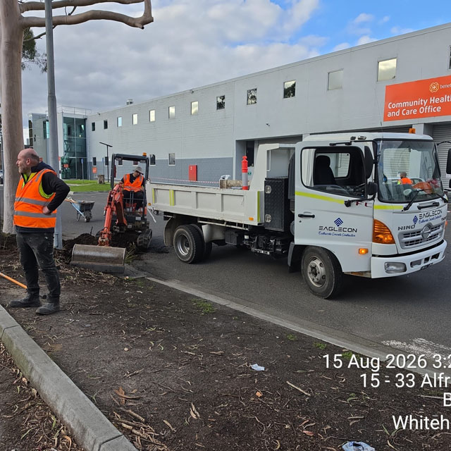 Worker in hi-vis beside a mini excavator and tipper truck on a garden strip