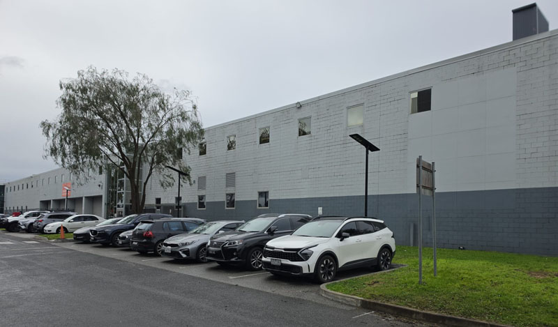 Cars parked along a long grey commercial building beside a lawn