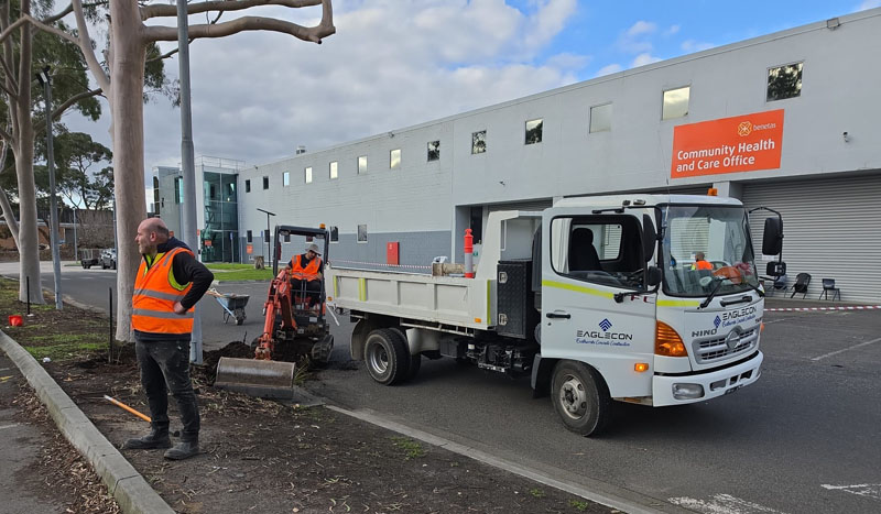 Worker in hi-vis beside a mini excavator and tipper truck outside a health care building