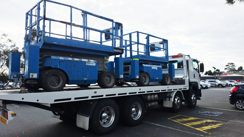 Tilt-tray truck carrying two blue scissor lifts in a car park