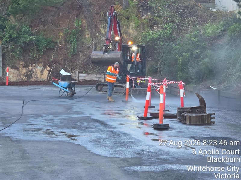Crew pressure-cleaning a concrete yard beside an excavator and safety barriers