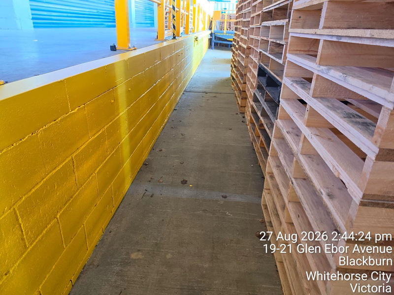 Concrete walkway between a yellow painted block wall and stacked timber pallets